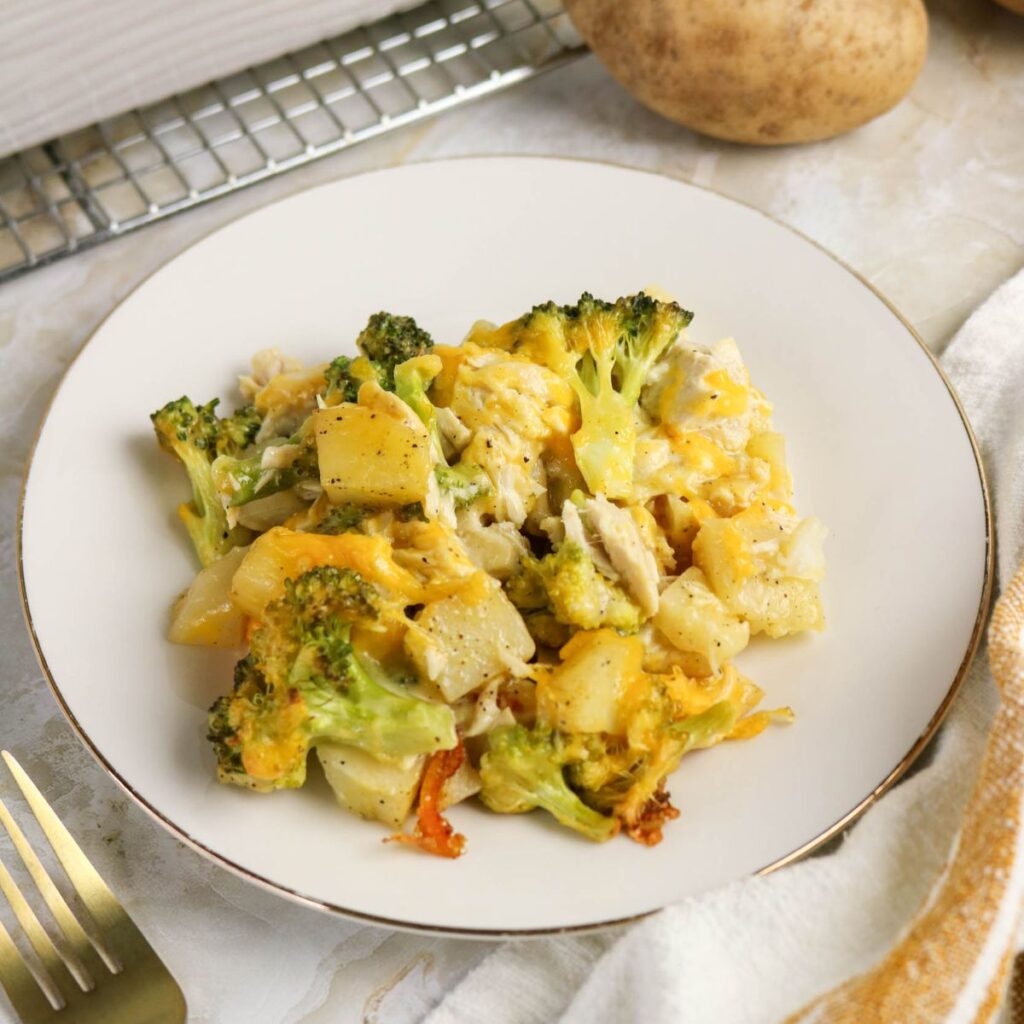 A serving of broccoli, potato, and cheese casserole on a white plate, set on a light marble surface with a fork and napkin nearby.