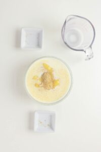A glass bowl with milk, sugar, and spices is on a white surface, surrounded by two small empty square dishes and a measuring cup containing liquid.
