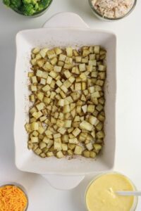 A baking dish filled with seasoned, diced potatoes, surrounded by bowls of shredded cheese, yellow sauce, chopped broccoli, and shredded chicken.