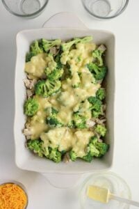 A white baking dish filled with broccoli florets, chicken pieces, and a creamy yellow sauce, surrounded by empty glass bowls and shredded cheese on a white surface.