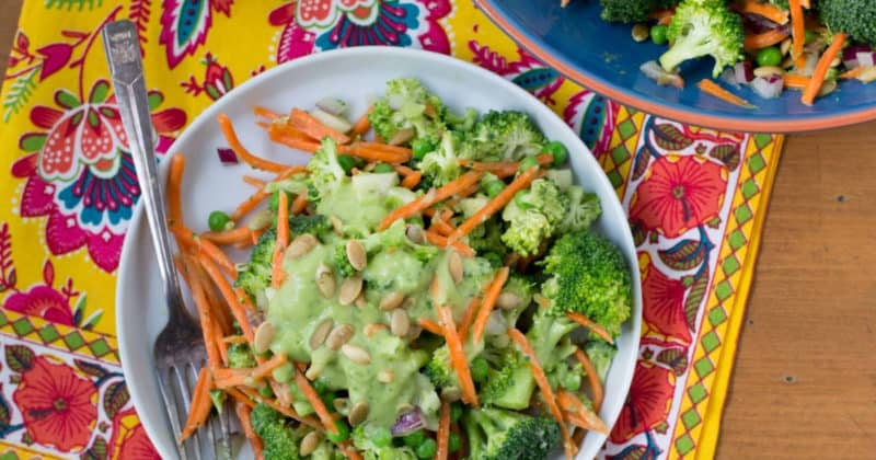 A plate of salad with broccoli, shredded carrots, peas, green dressing, and sunflower seeds sits on a colorful patterned tablecloth next to a fork and a serving bowl.