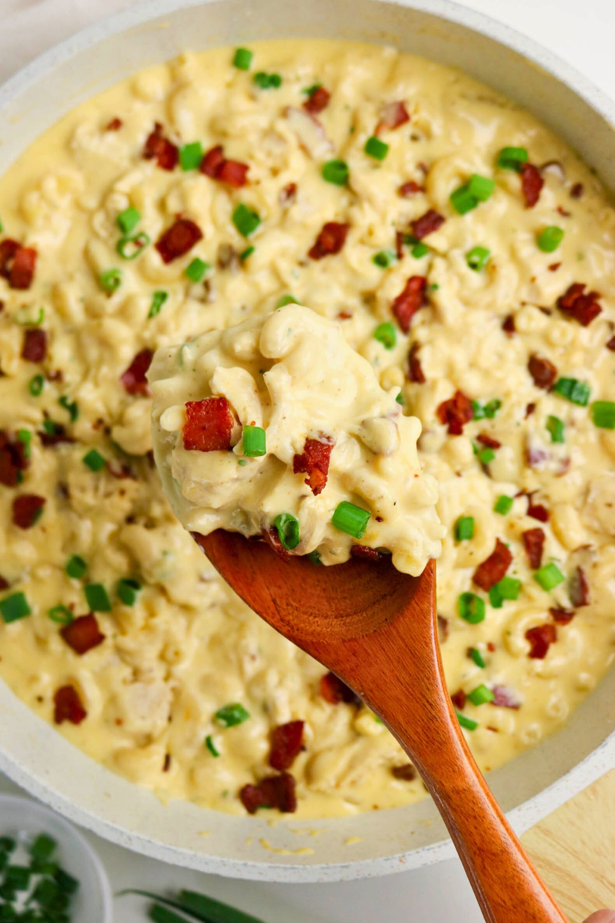 A wooden spoon holds a serving of creamy macaroni and cheese topped with bacon bits and chopped green onions above a pan of the same dish.