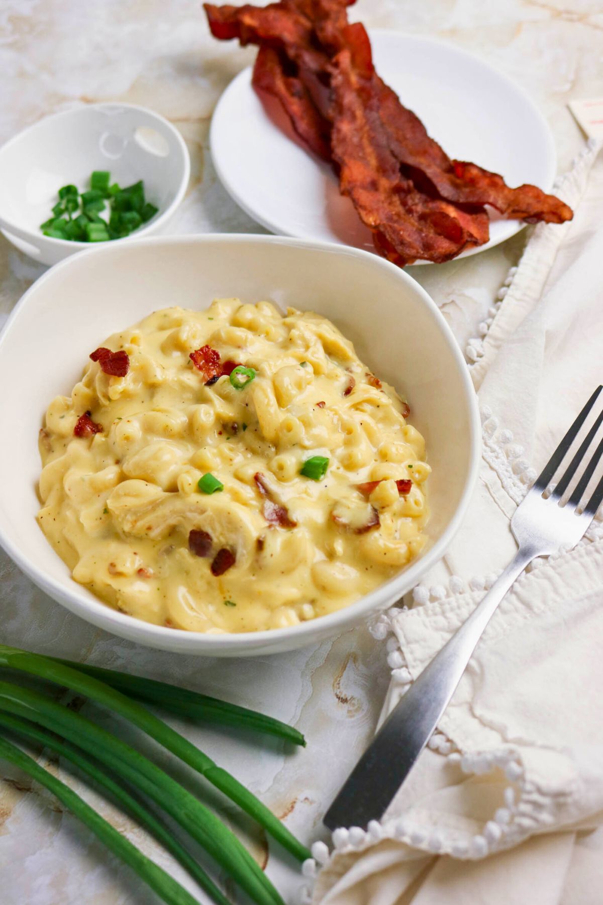 A bowl of macaroni and cheese topped with chopped green onions and bacon bits, with plates of bacon, green onions, and a fork nearby.