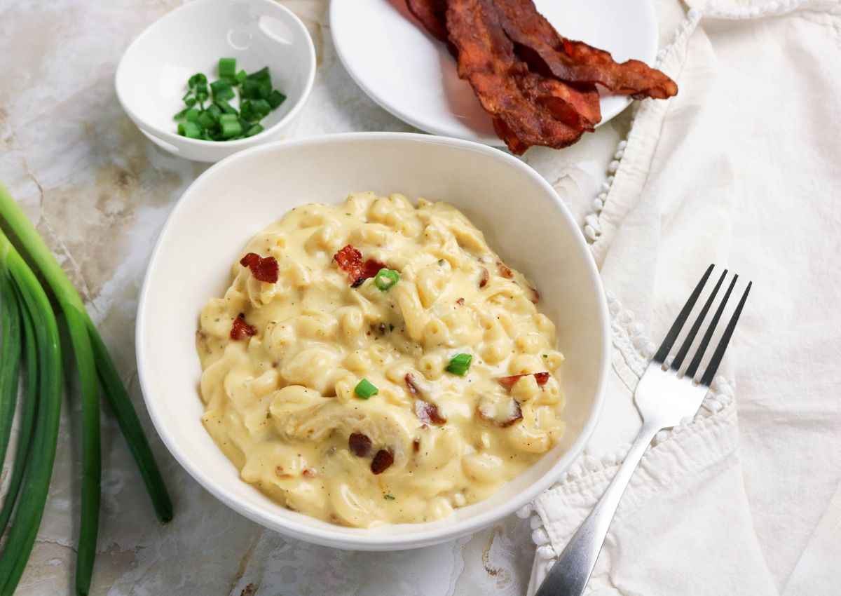 A bowl of creamy macaroni and cheese topped with bacon bits and green onions, with a plate of bacon, a small bowl of chopped green onions, and a fork on the side.