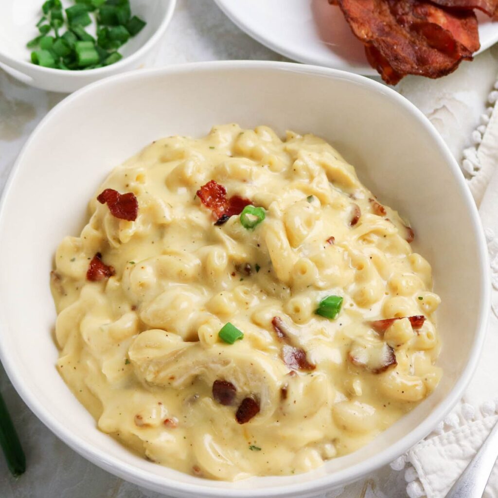 A bowl of creamy macaroni and cheese topped with bacon bits and chopped green onions, with a side dish of green onions and a plate of bacon in the background.
