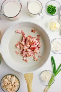 A white pan with chopped raw bacon in the center, surrounded by bowls containing milk, cream, butter, flour, green onions, cooked chicken, and seasonings.