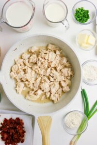 A skillet of cooked, chopped chicken surrounded by bowls of milk, cream, butter, flour, green onions, chopped bacon, and seasonings on a white surface.