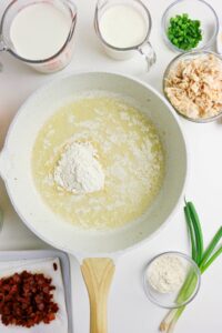 A skillet with melted butter and flour surrounded by small bowls of milk, green onions, shredded chicken, cooked bacon, and additional flour on a white surface.