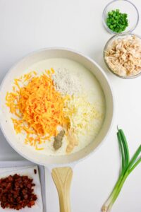 A skillet with milk, shredded cheddar and mozzarella cheese, flour, and seasonings. Nearby are bowls of cooked chicken, chopped green onions, and bacon bits.