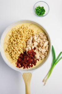 A skillet with macaroni, shredded chicken, chopped bacon, and cheese sauce, next to a bowl of sliced green onions and two whole green onions on a white background.