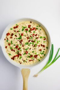 A skillet filled with creamy macaroni and cheese, topped with chopped bacon and green onions, sits on a white surface next to two fresh green onions.