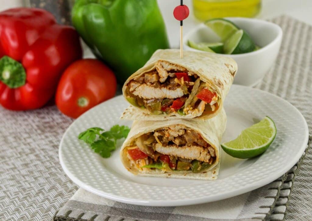 A sliced chicken fajita wrap with vegetables is served on a white plate with a lime wedge, with bell peppers, a tomato, and a bowl of lime wedges in the background.