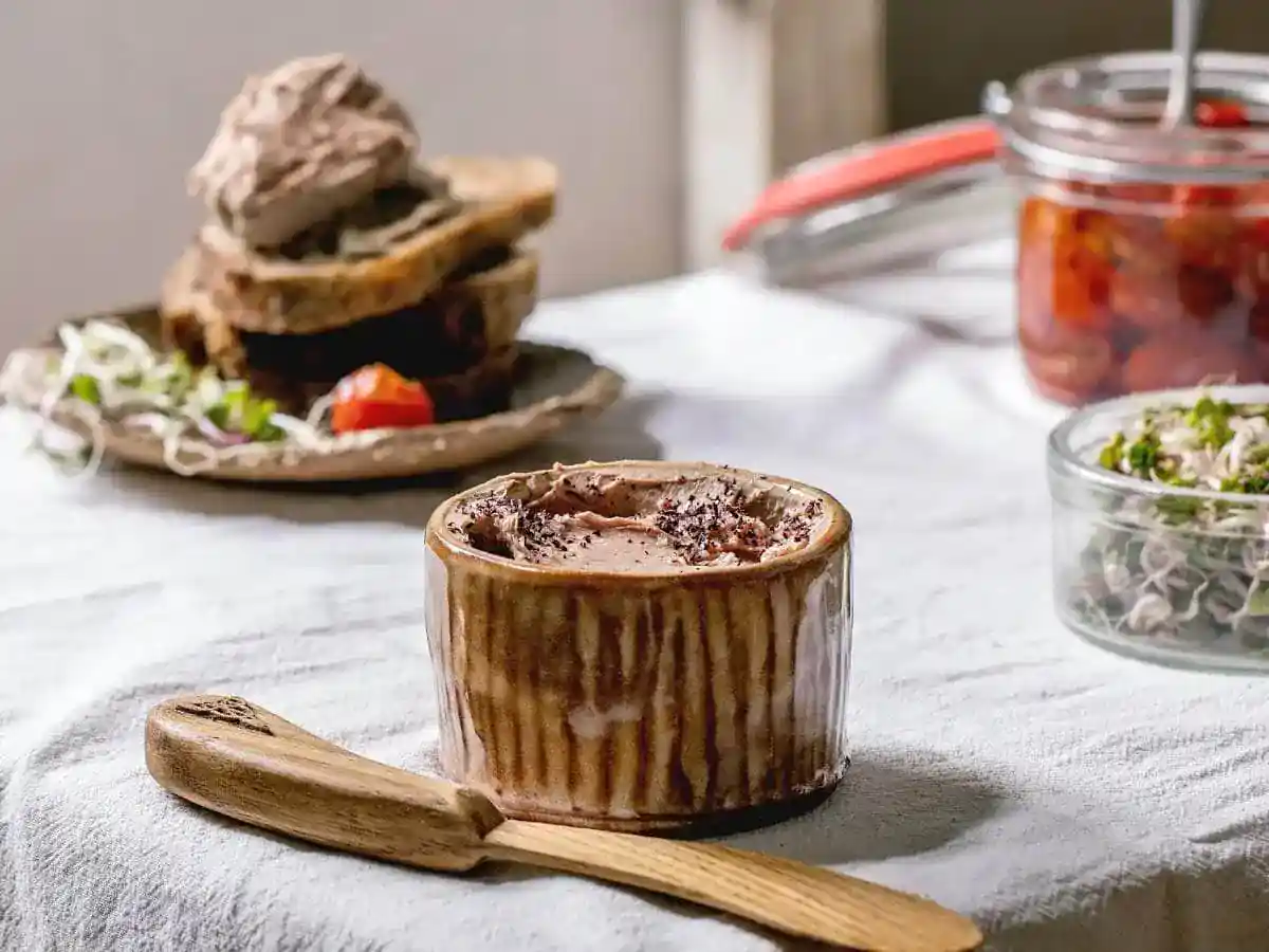 A ceramic dish filled with pâté sits on a table with a wooden spreader; bread, sprouts, and a jar of preserves are in the background.