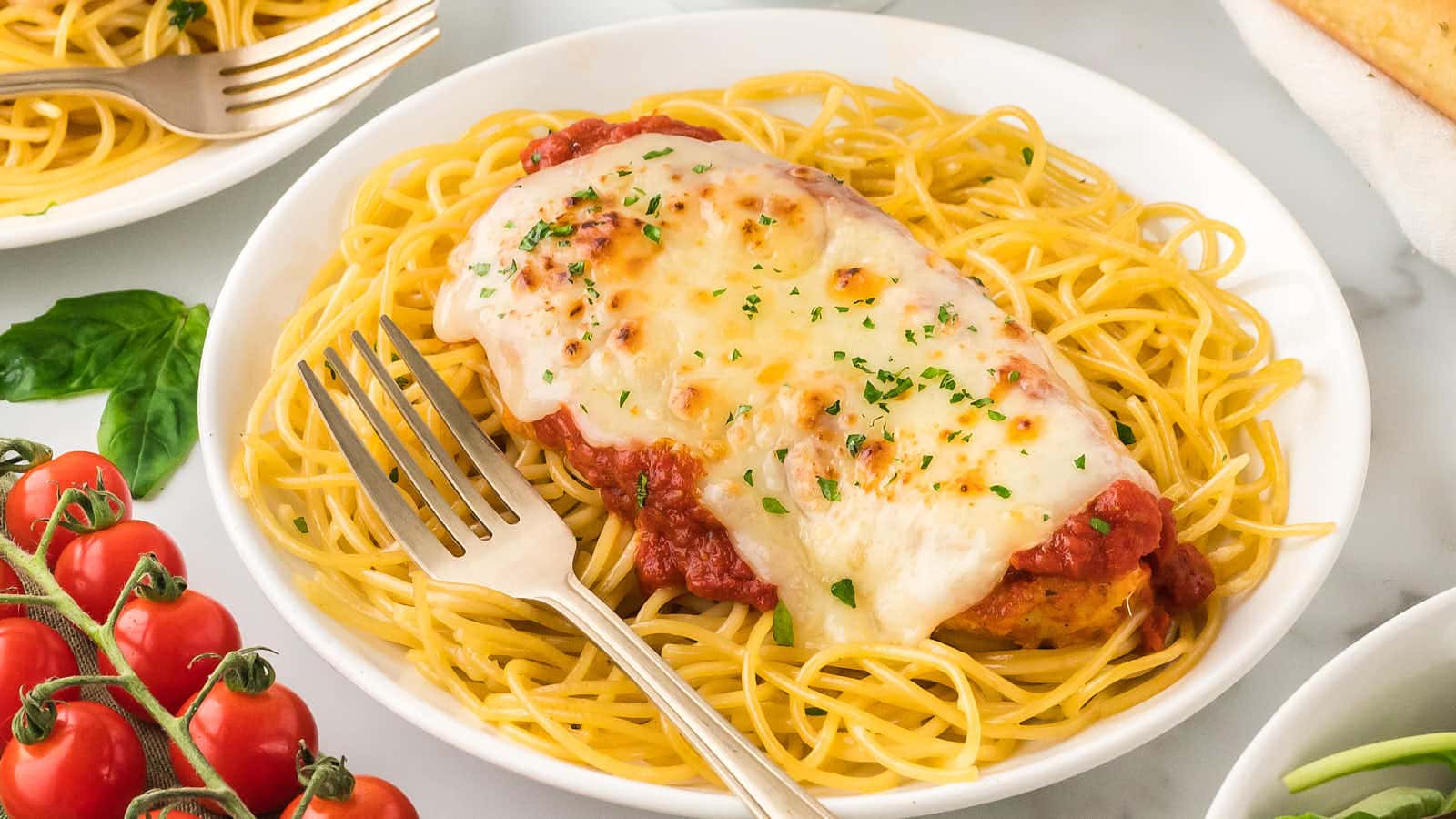 A plate of spaghetti topped with breaded chicken, marinara sauce, and melted cheese, with a fork on the side. Fresh tomatoes and basil are visible nearby.
