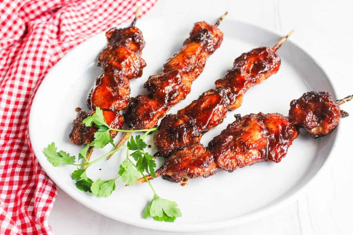 Four skewers of grilled, glazed meat are served on a white plate with a sprig of parsley, next to a red and white checkered cloth.