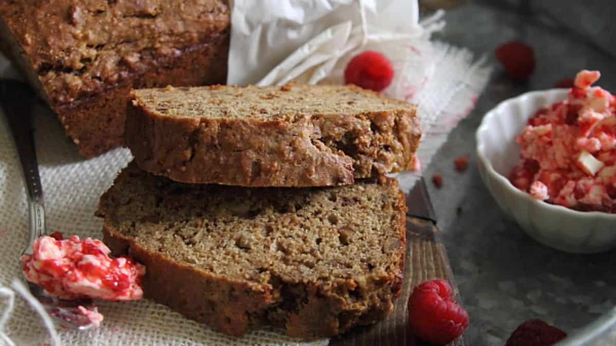 Two slices of banana bread are stacked on a cutting board next to a bowl of raspberry butter, with a loaf and fresh raspberries in the background.