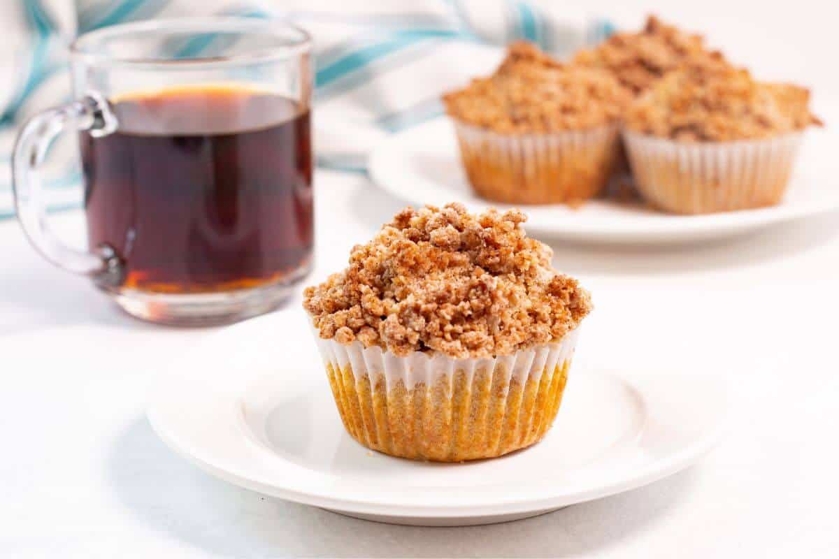 A crumb-topped muffin on a white plate sits next to a glass mug of black coffee, with more muffins blurred in the background.