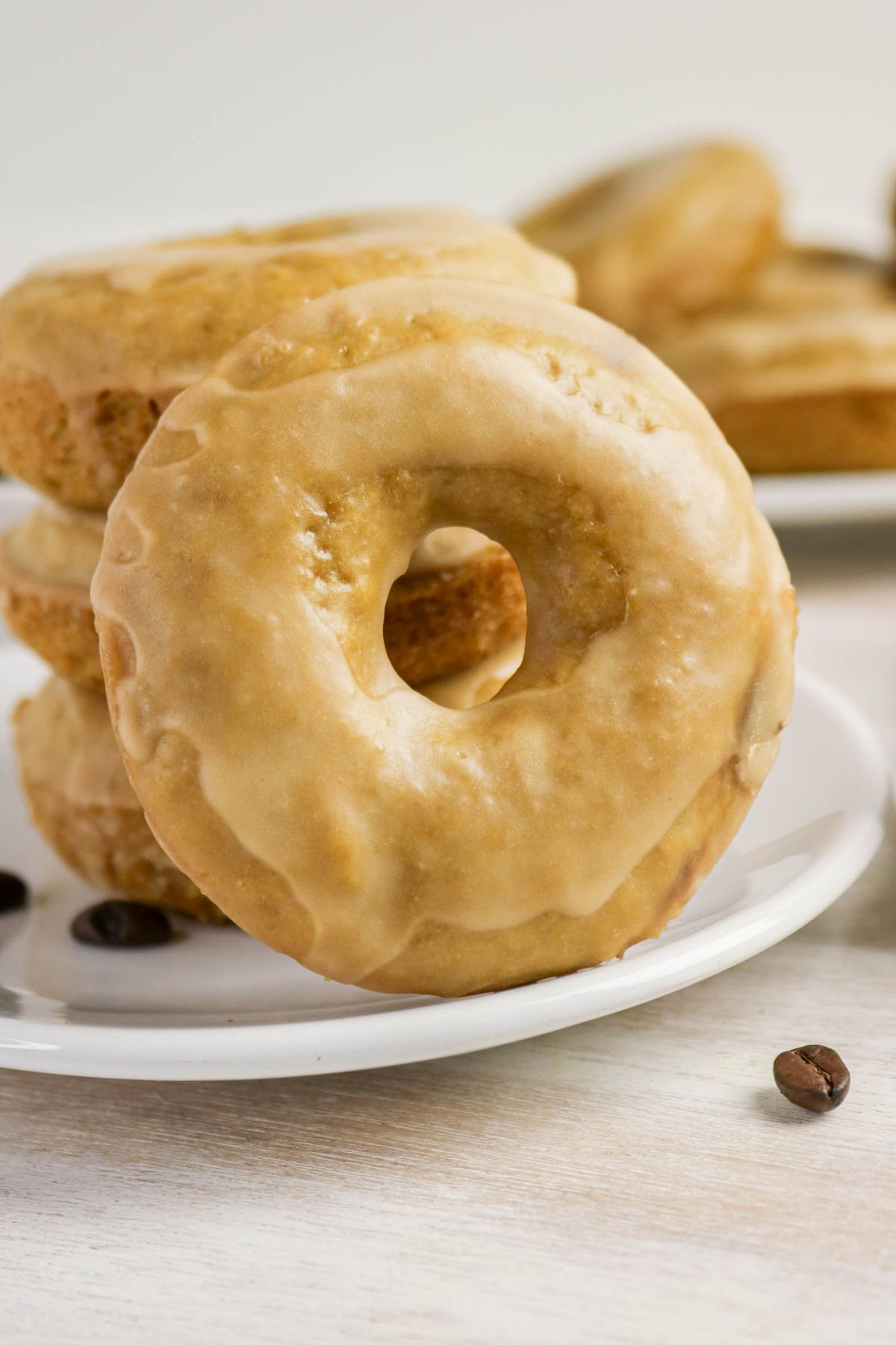 A plate of glazed coffee doughnuts is stacked, with one doughnut resting upright in front; coffee beans are scattered on the table nearby.