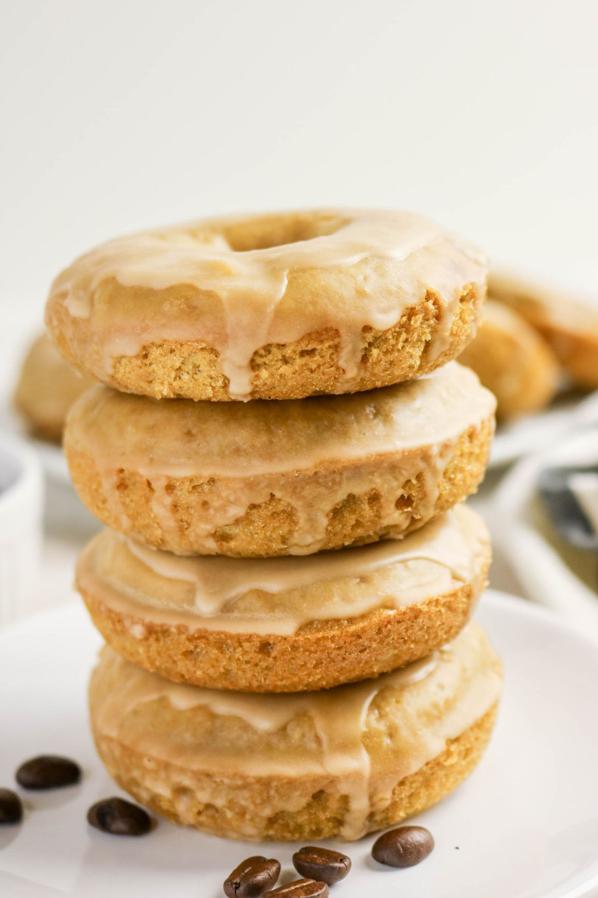 Three coffee doughnuts are stacked on a white plate with a few coffee beans scattered nearby.