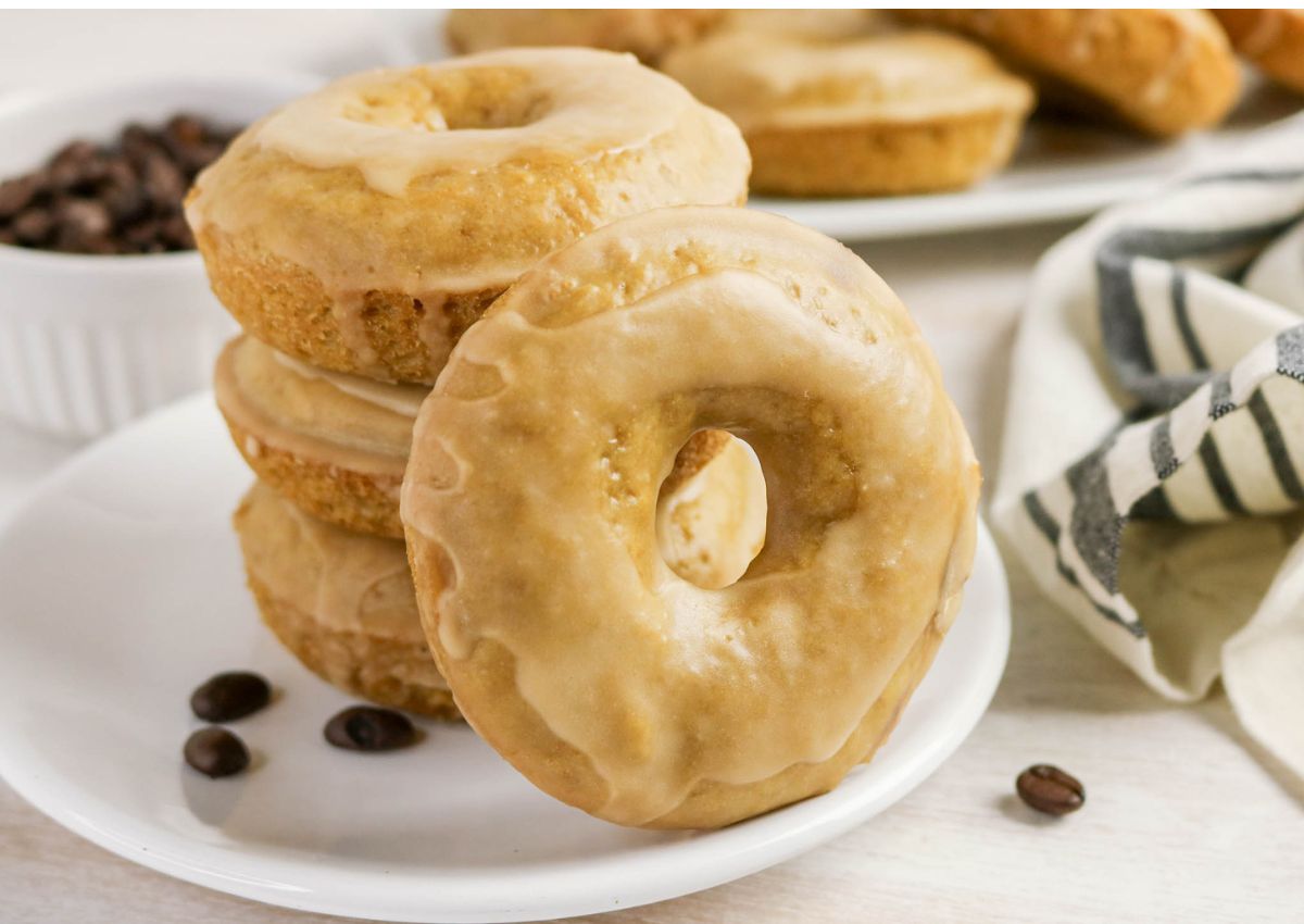 A close-up of four Coffee Doughnuts stacked on a white plate, with a few coffee beans scattered nearby and a bowl of beans in the background.