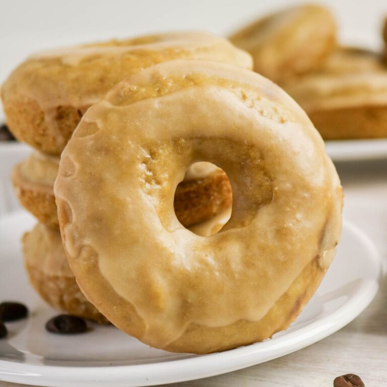 A stack of glazed coffee doughnuts sits on a white plate, with a few coffee beans scattered nearby.