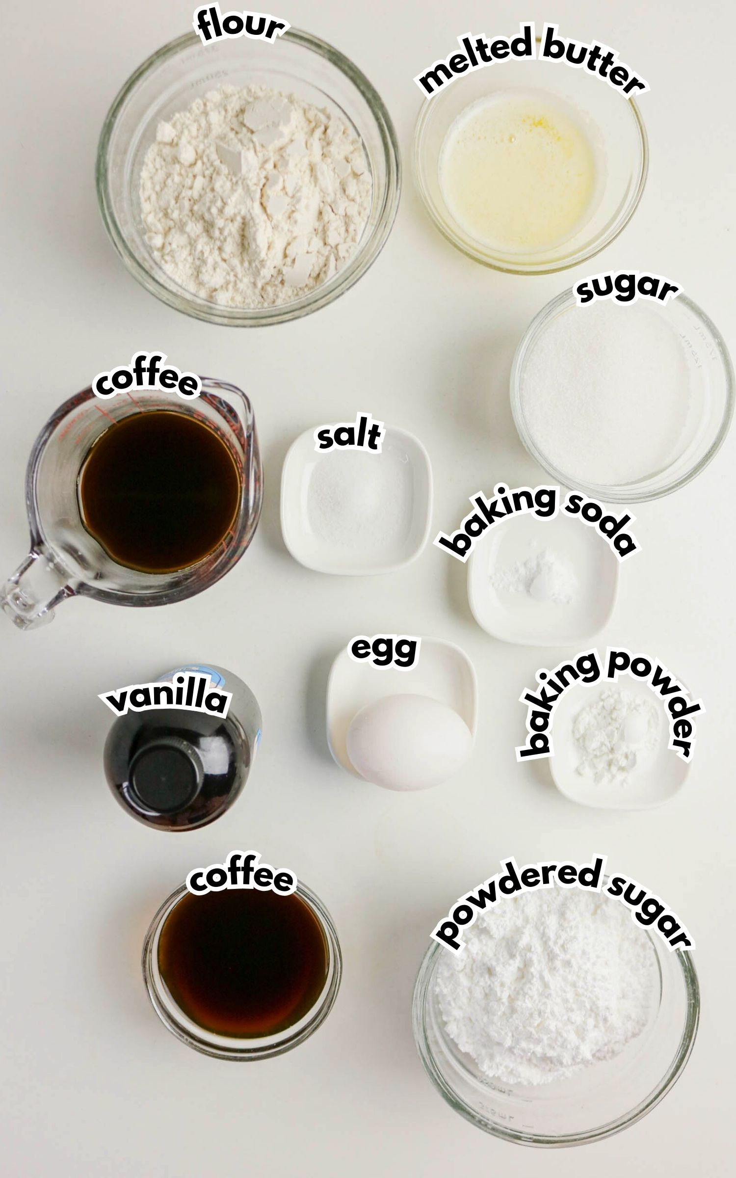 Overhead view of labeled baking ingredients in bowls for Coffee Doughnuts, including flour, melted butter, sugar, two portions of coffee, vanilla, salt, egg, baking soda, baking powder, and powdered sugar.