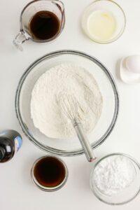 Overhead view of baking ingredients for Coffee Doughnuts, featuring flour in a bowl with a whisk, coffee, melted butter, vanilla extract, powdered sugar, and an egg on a white surface.