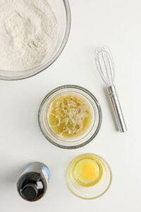A bowl of flour, a whisk, a bottle of vanilla extract, a small bowl with a raw egg, and a bowl with melted butter are arranged on a white surface—perfect ingredients for making delicious Coffee Doughnuts.