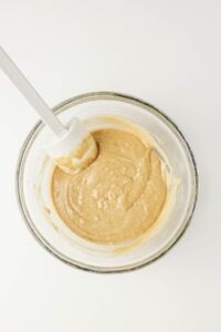 A glass bowl filled with light brown Coffee Doughnuts batter and a white spatula resting inside, set on a white surface.
