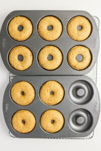 Two six-cavity metal donut pans, each holding six freshly baked coffee doughnuts, are placed on a wire cooling rack against a white background.