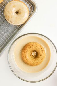 A baked coffee doughnut is being dipped in glaze inside a glass bowl, with a finished glazed donut resting on a wire rack nearby.