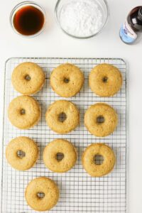 A cooling rack with ten plain baked coffee doughnuts sits next to bowls of powdered sugar, dark liquid, and a bottle of vanilla extract on a white surface.