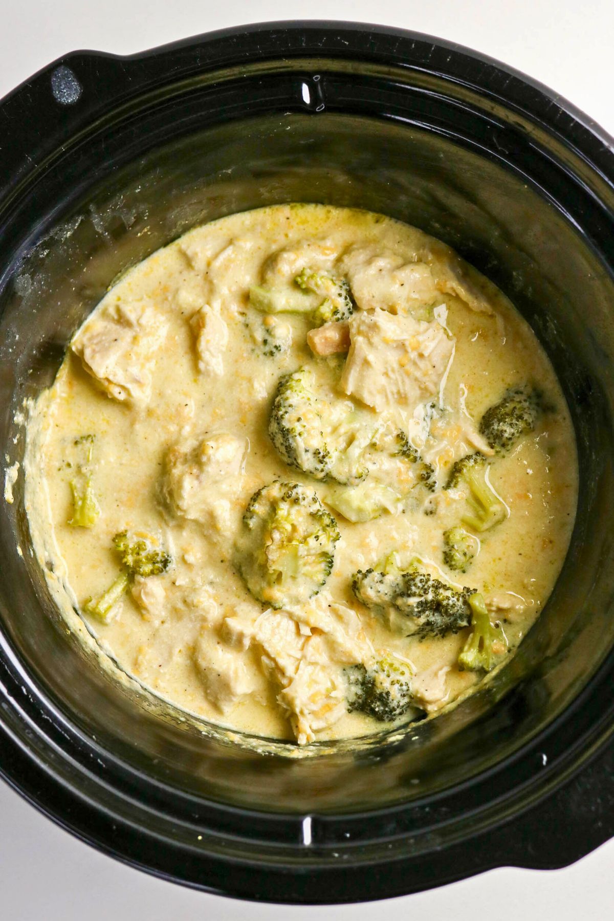 Chicken, broccoli, and creamy sauce cooked together in a black slow cooker, viewed from above.