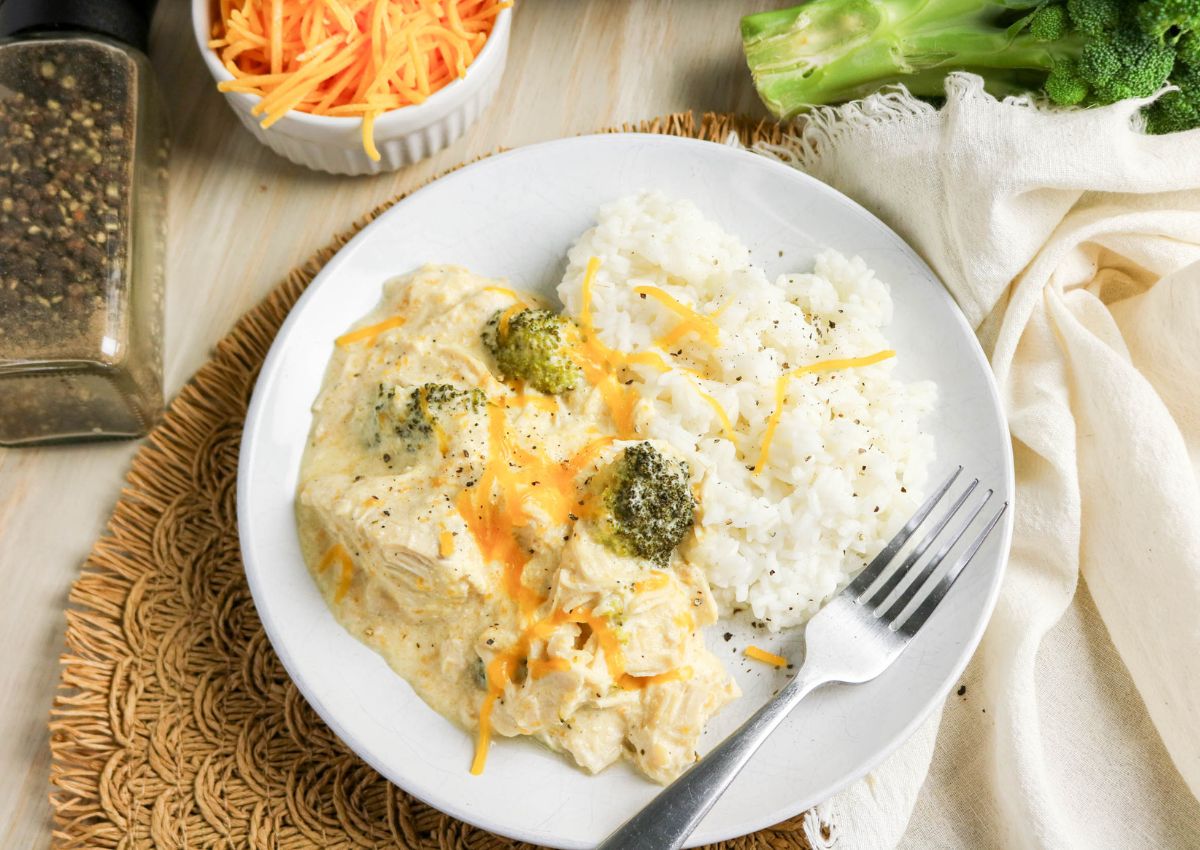 A white plate with white rice and creamy chicken broccoli casserole, topped with shredded cheese and black pepper, with a fork on the side.
