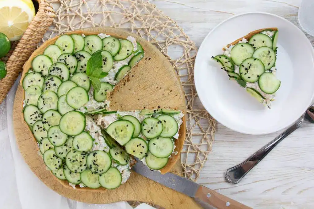 A round savory tart topped with sliced cucumbers, black sesame seeds, and herbs sits on a wooden board with one slice served on a white plate beside it.