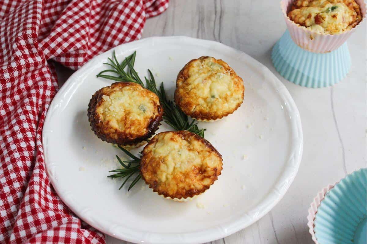 Four savory muffins are arranged on a white plate with rosemary sprigs, next to a red checkered cloth and additional muffins in paper liners.