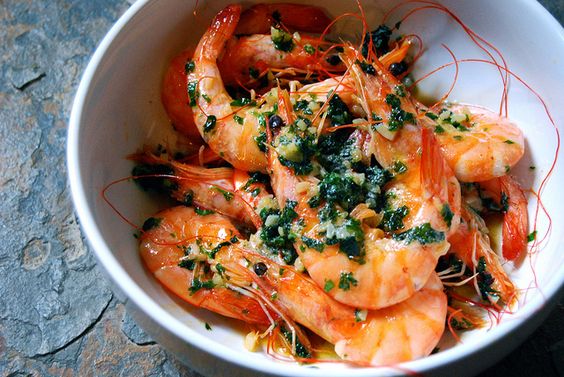 A bowl of cooked shrimp garnished with chopped herbs and garlic in a light sauce, placed on a stone surface.