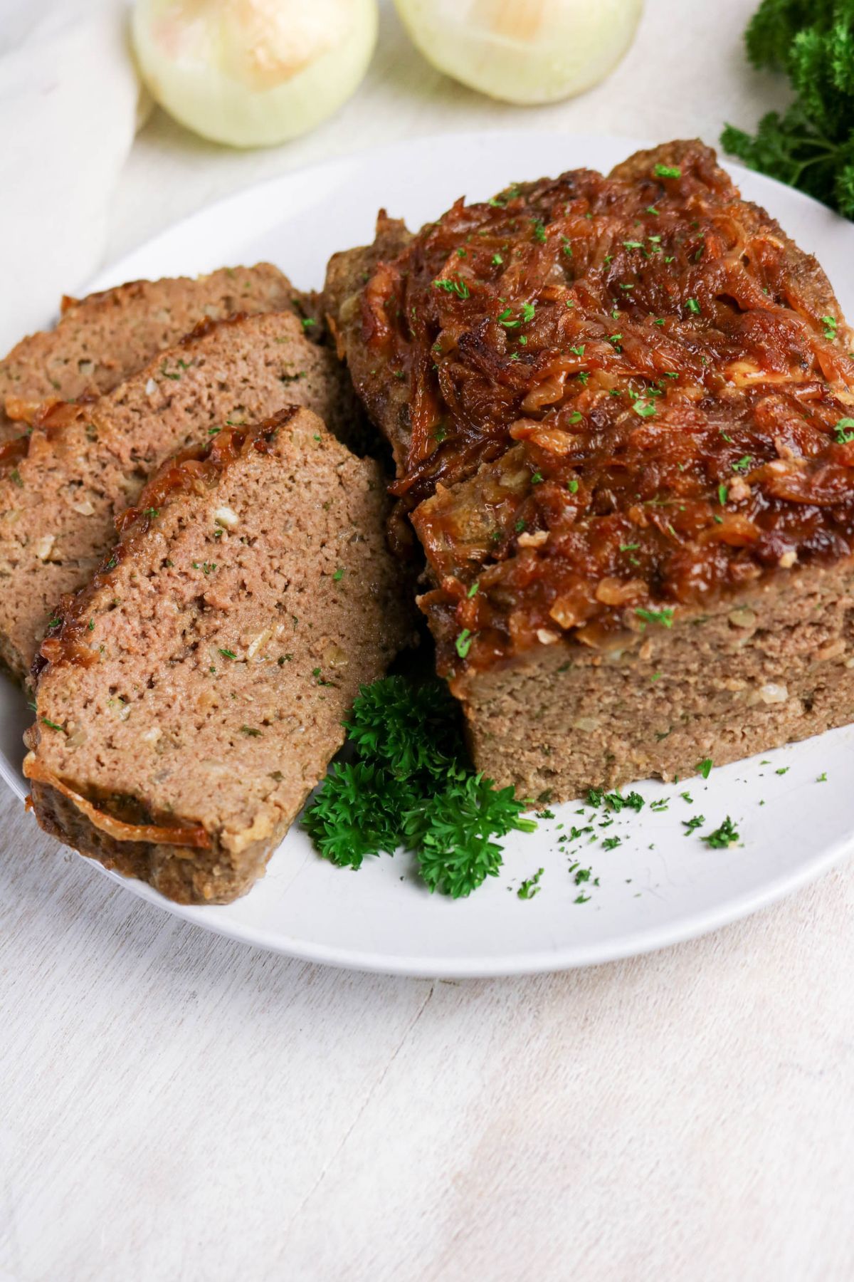 A plate of sliced meatloaf topped with caramelized onions, garnished with fresh parsley, on a white surface with onions in the background.
