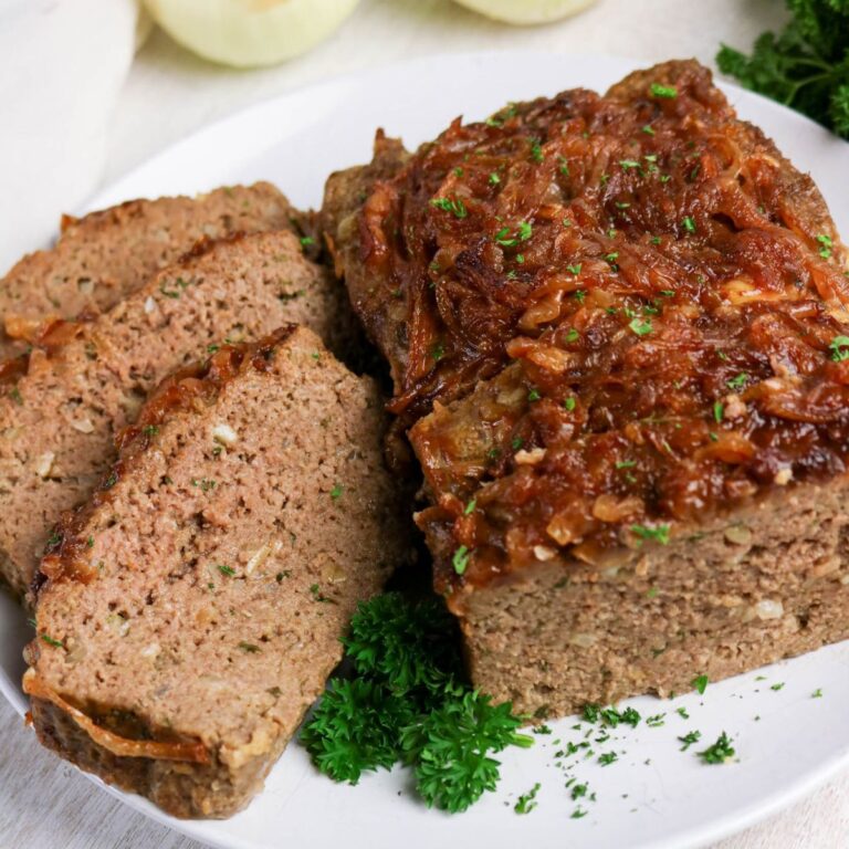 A sliced meatloaf topped with caramelized onions is served on a white plate, garnished with fresh parsley.