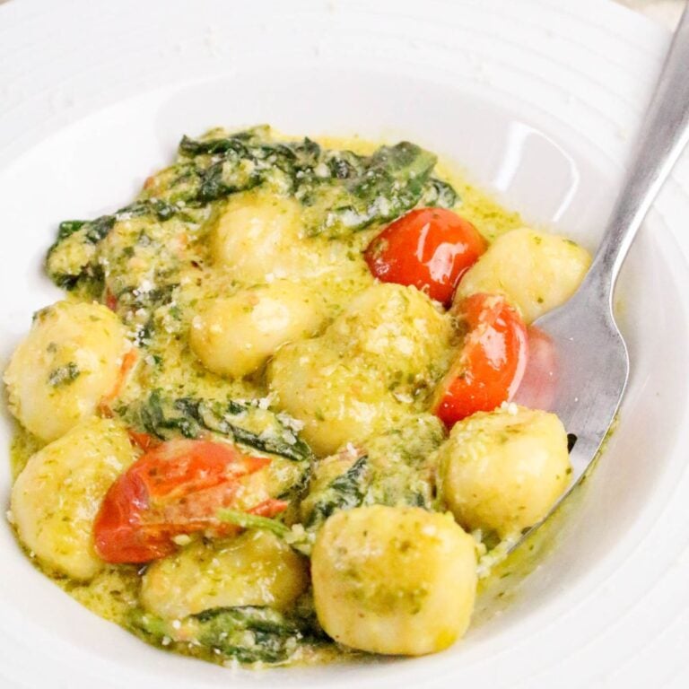 A white plate of gnocchi with pesto sauce, spinach, cherry tomatoes, and grated cheese, with a fork on the side.