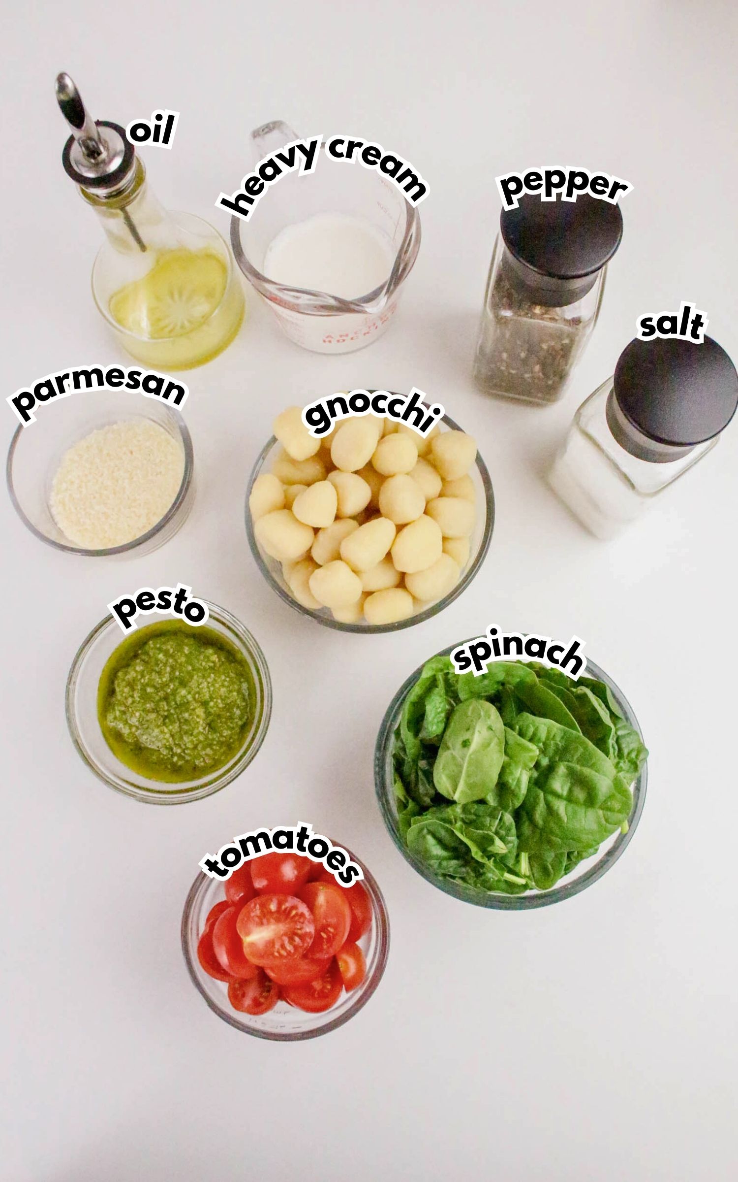 Ingredients for a gnocchi dish arranged on a white surface: oil, heavy cream, pepper, salt, parmesan, gnocchi, pesto, spinach, and sliced tomatoes in separate containers.