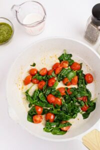 A white pan with sautéed spinach and cherry tomatoes, surrounded by a measuring cup of milk, a jar of pesto, and a pepper grinder on a white surface.