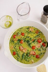 A white pan containing a creamy sauce with spinach, cherry tomatoes, and sausage sits on a white surface beside a glass measuring cup, an empty bowl, and a pepper shaker.