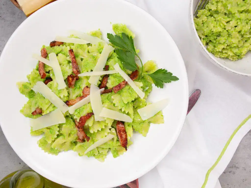 A white plate of farfalle pasta with green pea sauce, topped with bacon strips, Parmesan shavings, and a sprig of parsley. A bowl of extra green pea sauce is beside the plate.