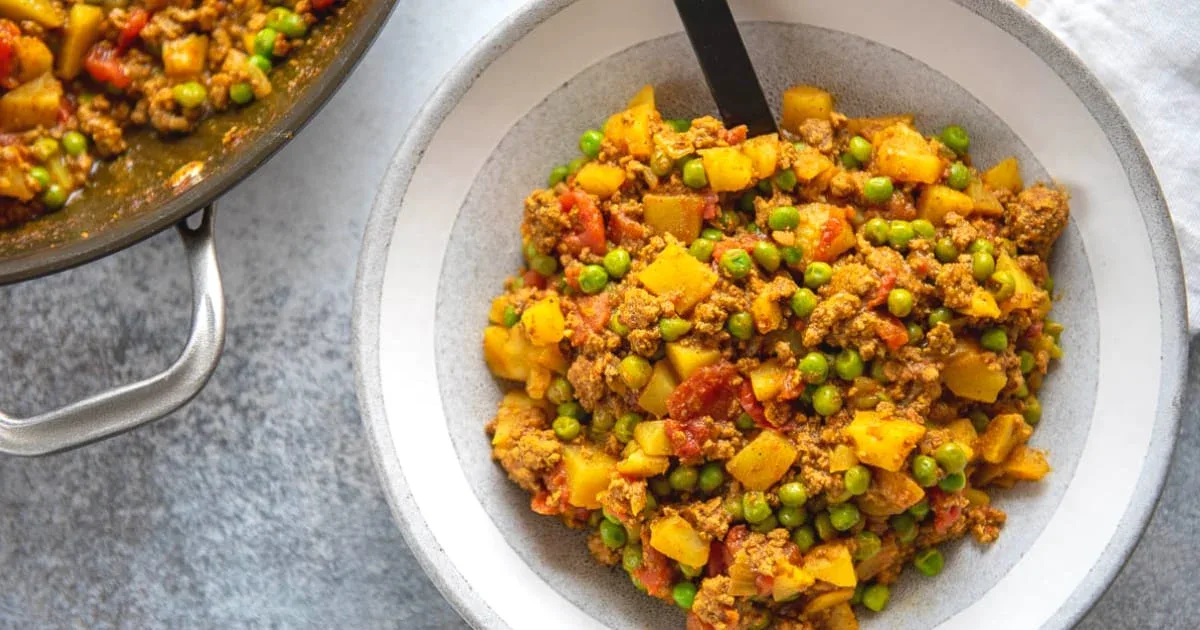 A bowl of ground meat curry with potatoes, peas, and tomatoes, served with a spoon on a white plate; a pot with more curry is partially visible.