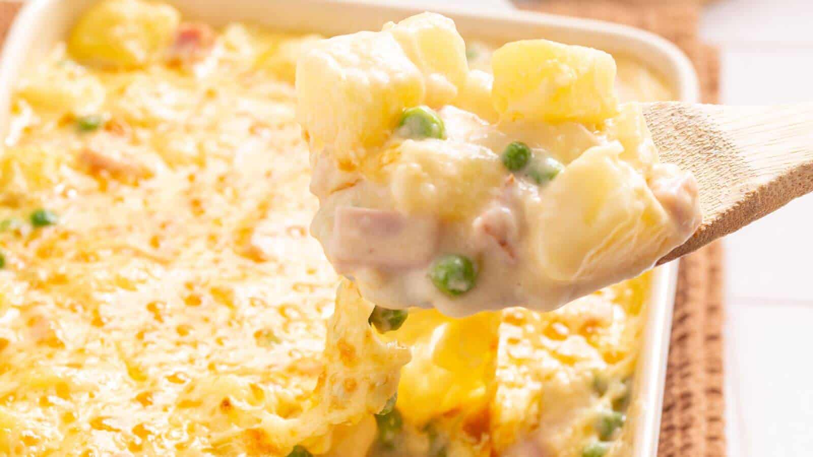 A wooden spoon lifts a portion of creamy baked casserole with peas, diced ham, and melted cheese from a baking dish.