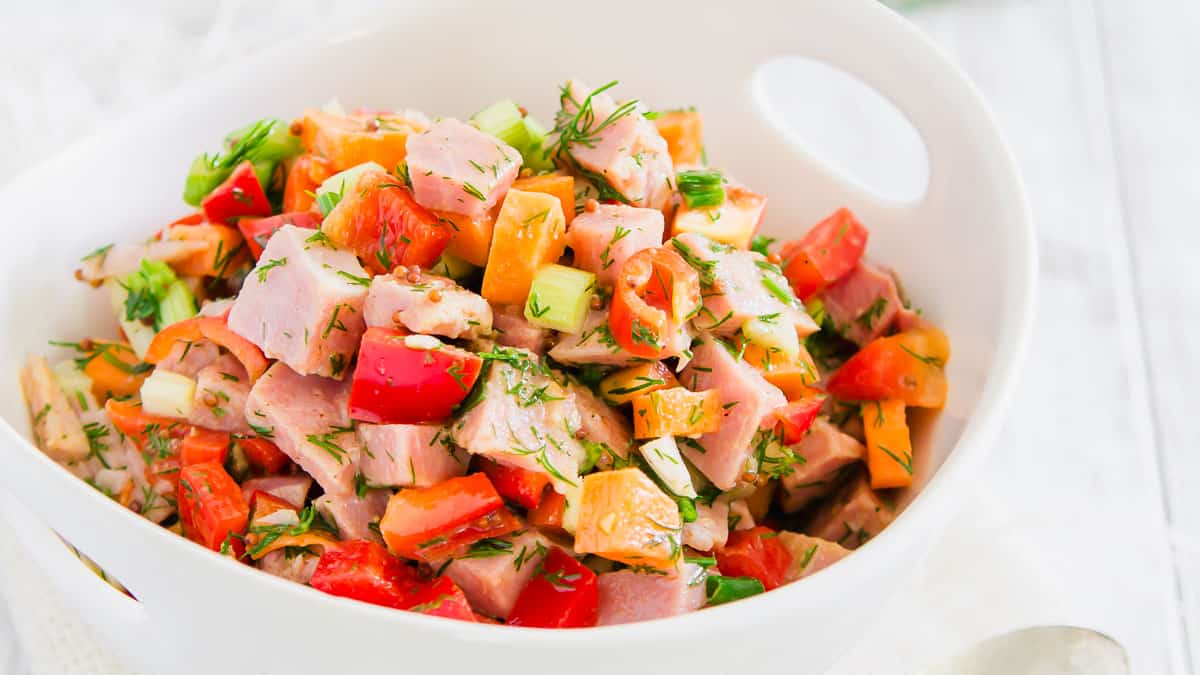 A white bowl filled with a chopped salad containing ham, red and orange bell peppers, green onions, and fresh dill.