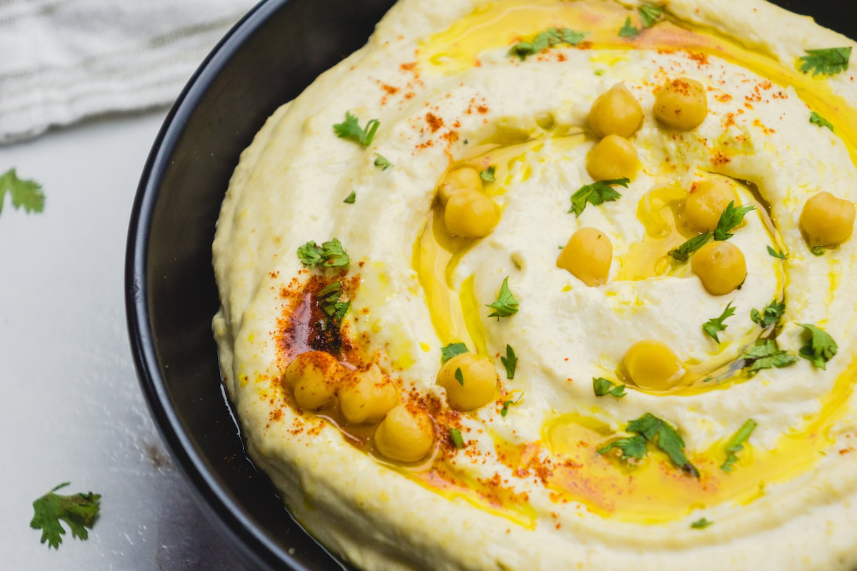 A black bowl filled with hummus, topped with whole chickpeas, olive oil, paprika, and chopped parsley.