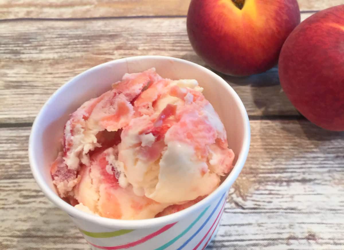 A cup of peach ice cream sits on a wooden surface with two whole peaches in the background.