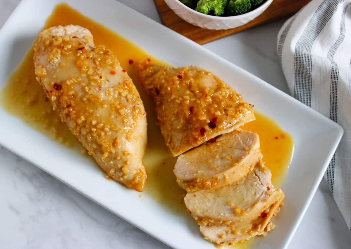 Two pieces of cooked chicken breast with a garlic and herb sauce, one cut into slices, served on a white rectangular plate.